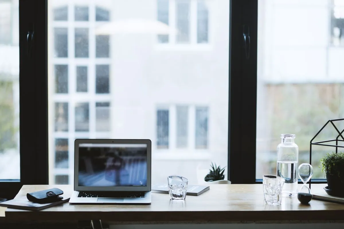 En laptop på et trebord ved et stort vindu, med et vannglass og små planter ved siden av
